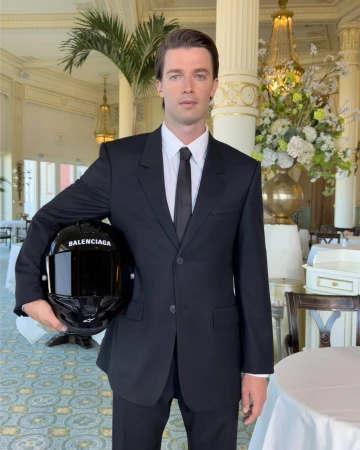 Man wearing a black suit and white shirt holding a branded black motorcycle helmet inside a hotel interior