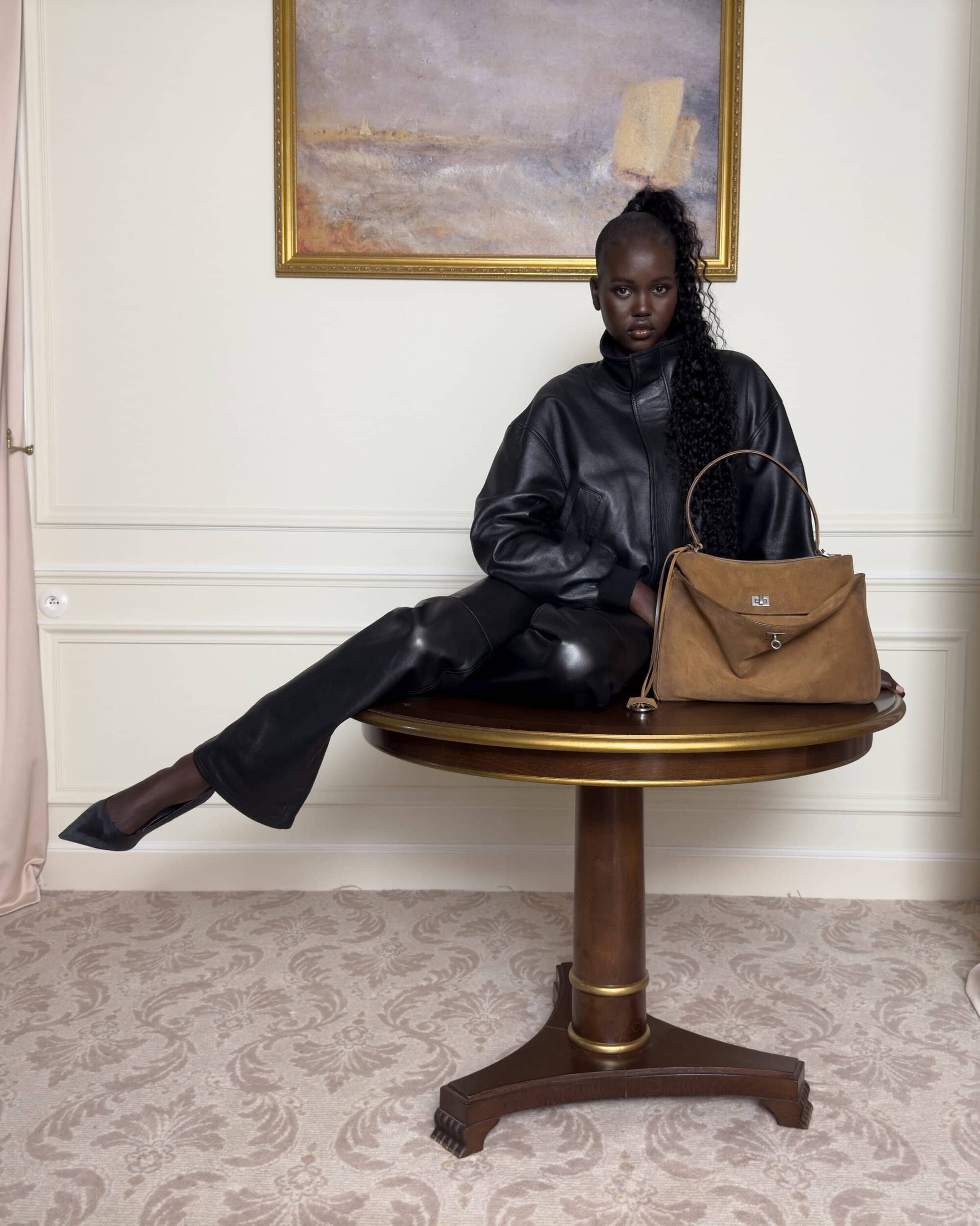 Model wearing black leather jacket seated on a round wooden table next to a brown suede handbag