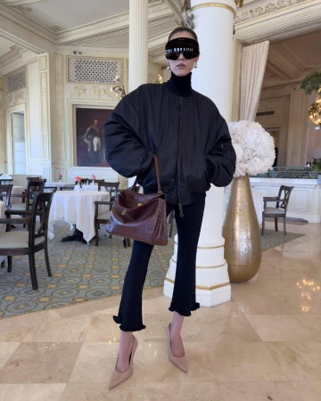 Model in oversized black bomber jacket and dark sunglasses holding brown leather handbag in ornate hotel interior