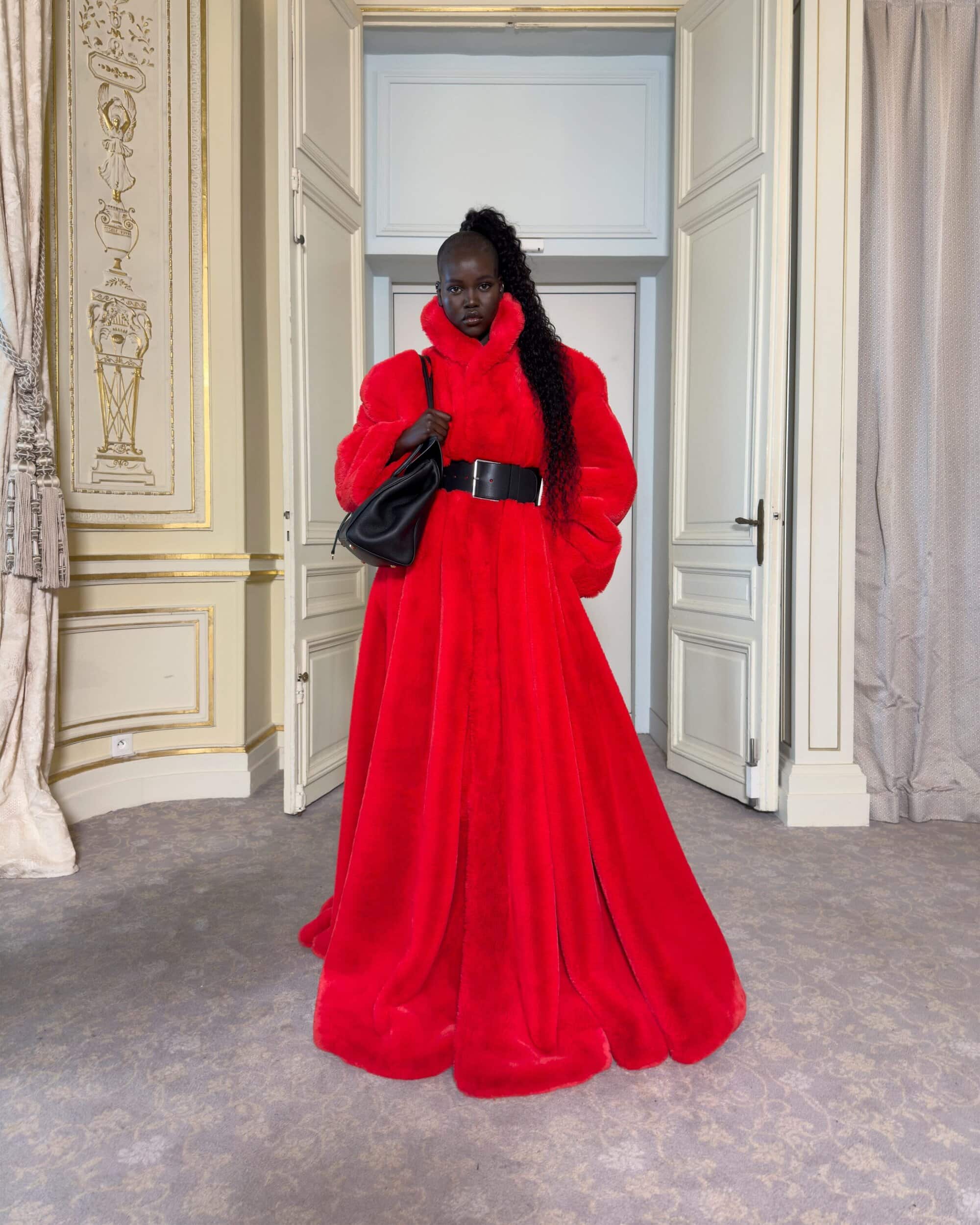 Model in voluminous red faux fur coat holding a black handbag inside an ornate room