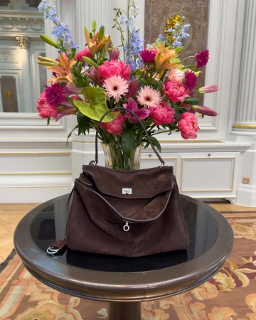 Dark brown suede handbag resting on a round black table in front of a large vase overflowing with colorful floral arrangements.