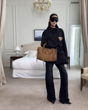 Model in black outerwear and visor sunglasses holding a tan leather bag inside an elegant hotel room