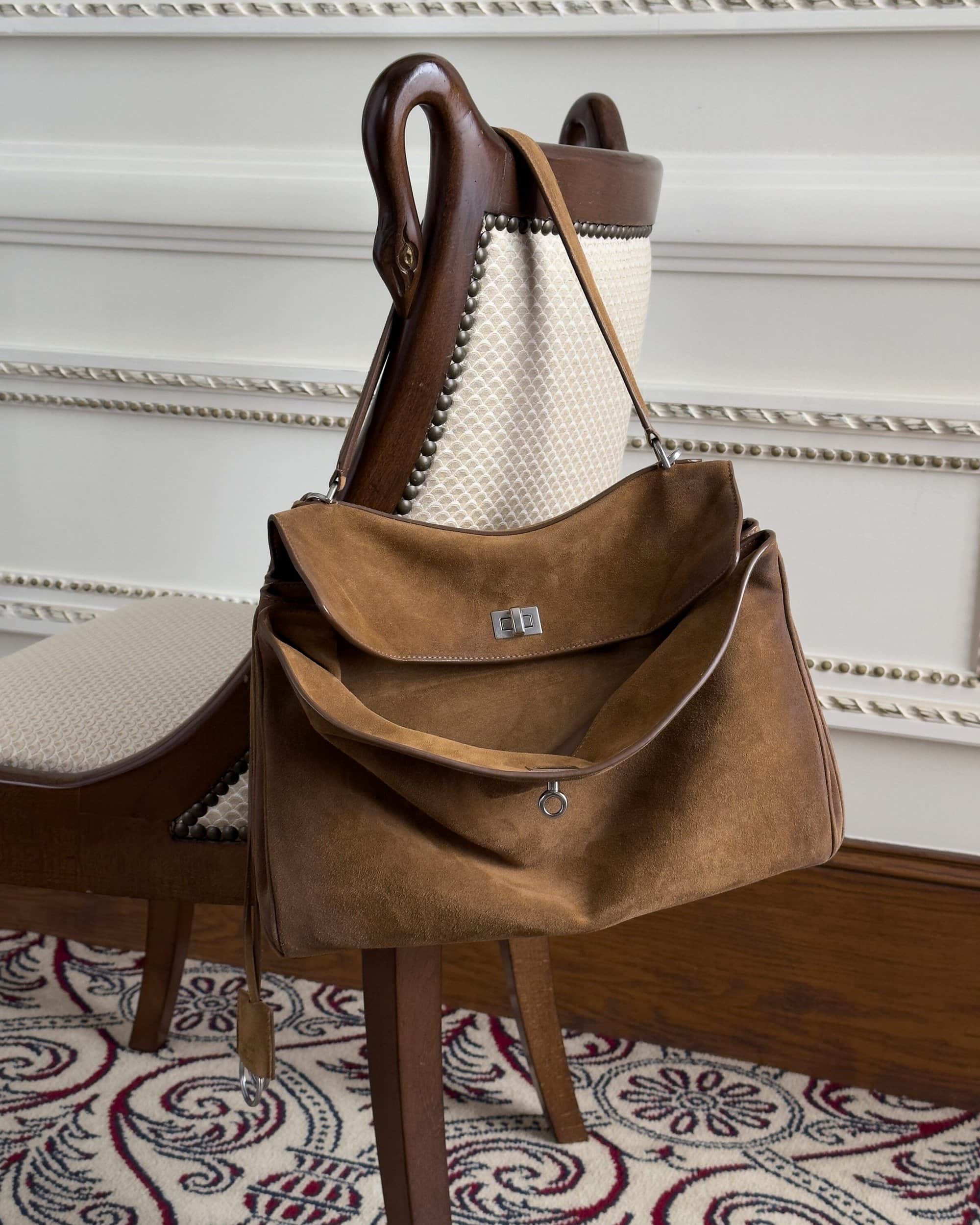 Brown suede shoulder bag draped over the back of an antique wooden carved chair in a classic room