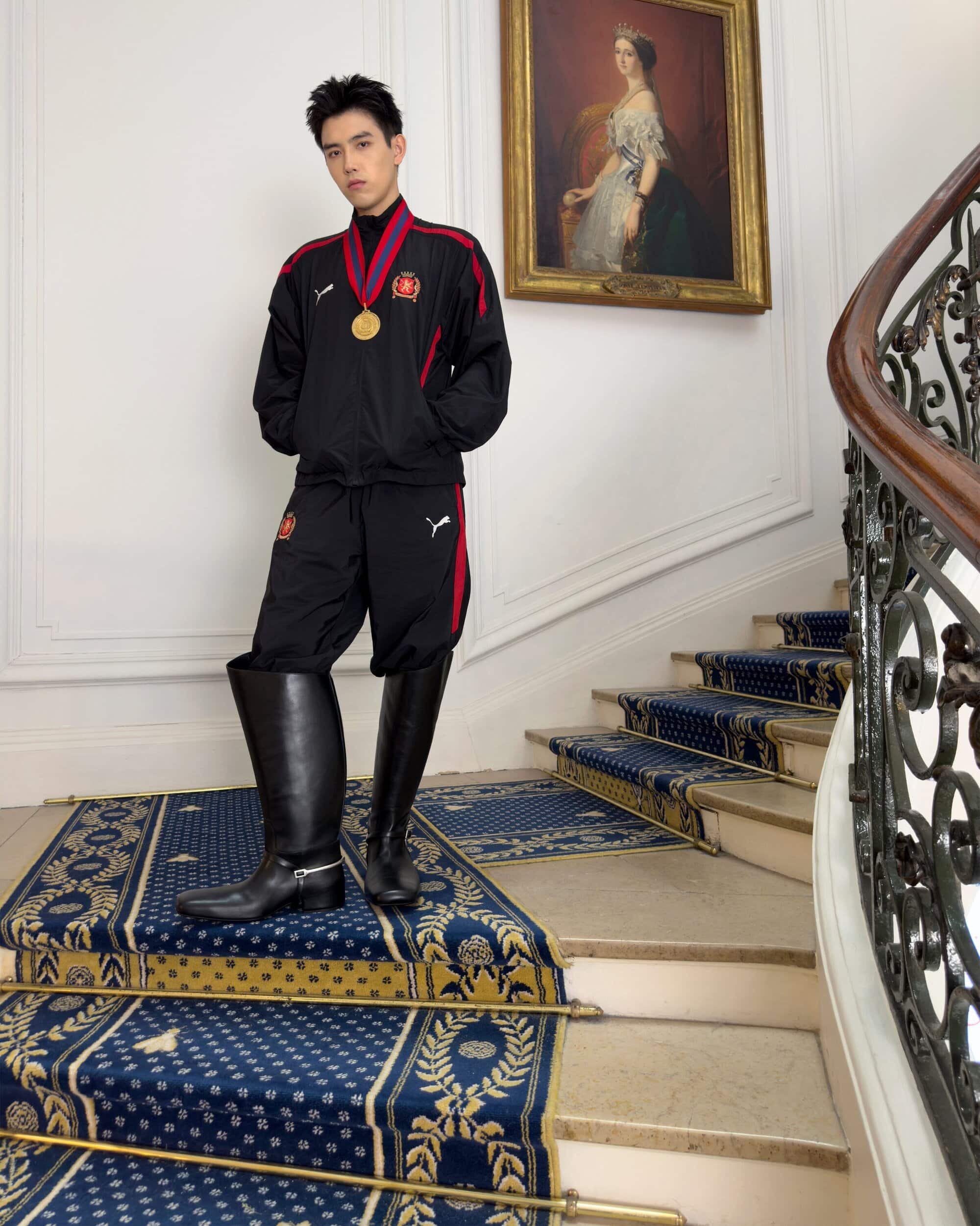 Man wearing a black tracksuit and tall leather boots standing on a grand carpeted staircase beneath a framed portrait