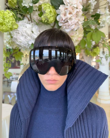 Model wearing dark reflective oversized sunglasses and a high-collared navy blue ribbed sweater against a lush floral background