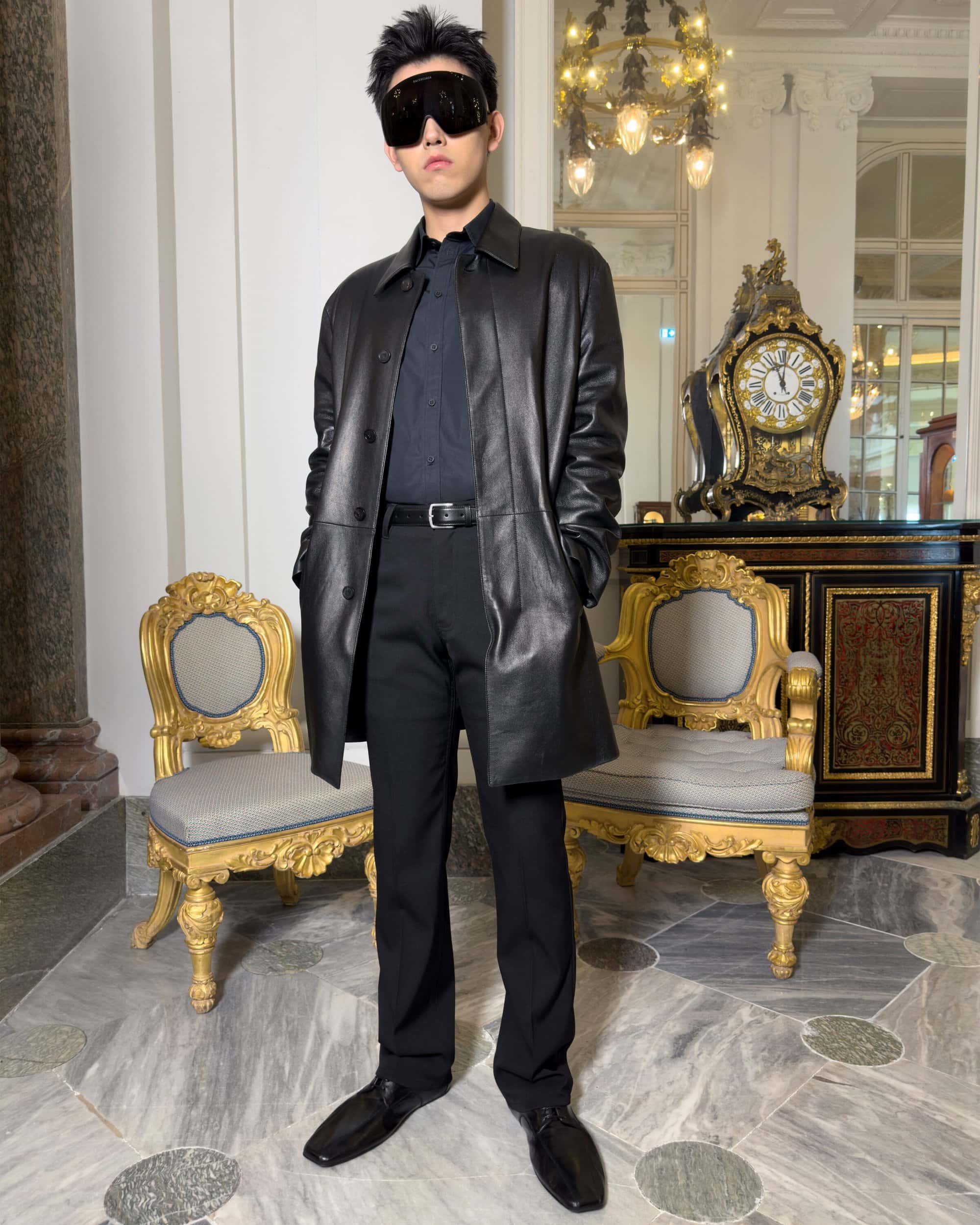 Man wearing a black leather coat, dark shirt, trousers, and sunglasses standing in an ornate room with gilded chairs.