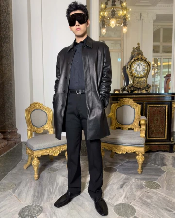 Man wearing a black leather coat, dark shirt, trousers, and sunglasses standing in an ornate room with gilded chairs.