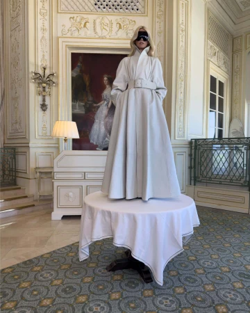 Model standing on a draped table wearing a long belted white coat and oversized sunglasses inside a luxurious hotel lobby