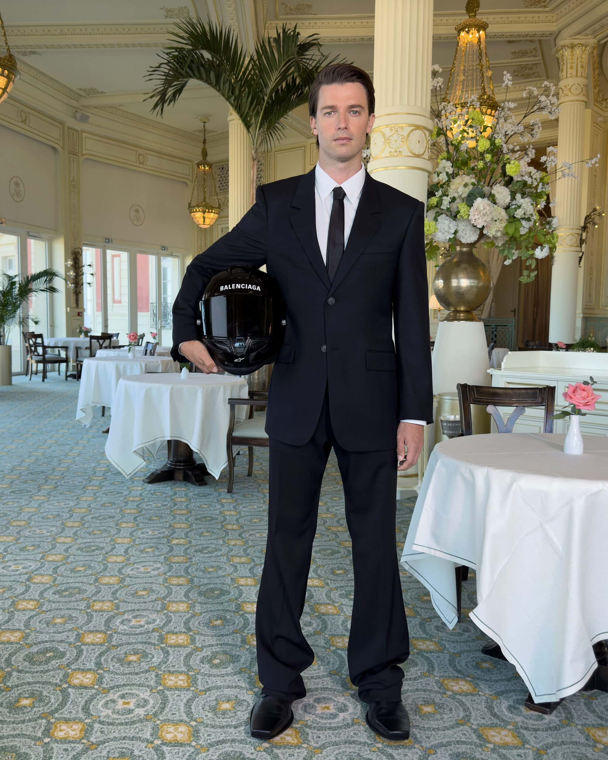 Man in a black suit holding a motorcycle helmet inside a hotel dining room for the Balenciaga Winter 2025 campaign.