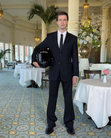 Man in a black suit holding a motorcycle helmet inside a hotel dining room for the Balenciaga Winter 2025 campaign.