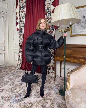 Isabelle Huppert in a black puffer jacket holding a handbag next to a lamp in a hotel room