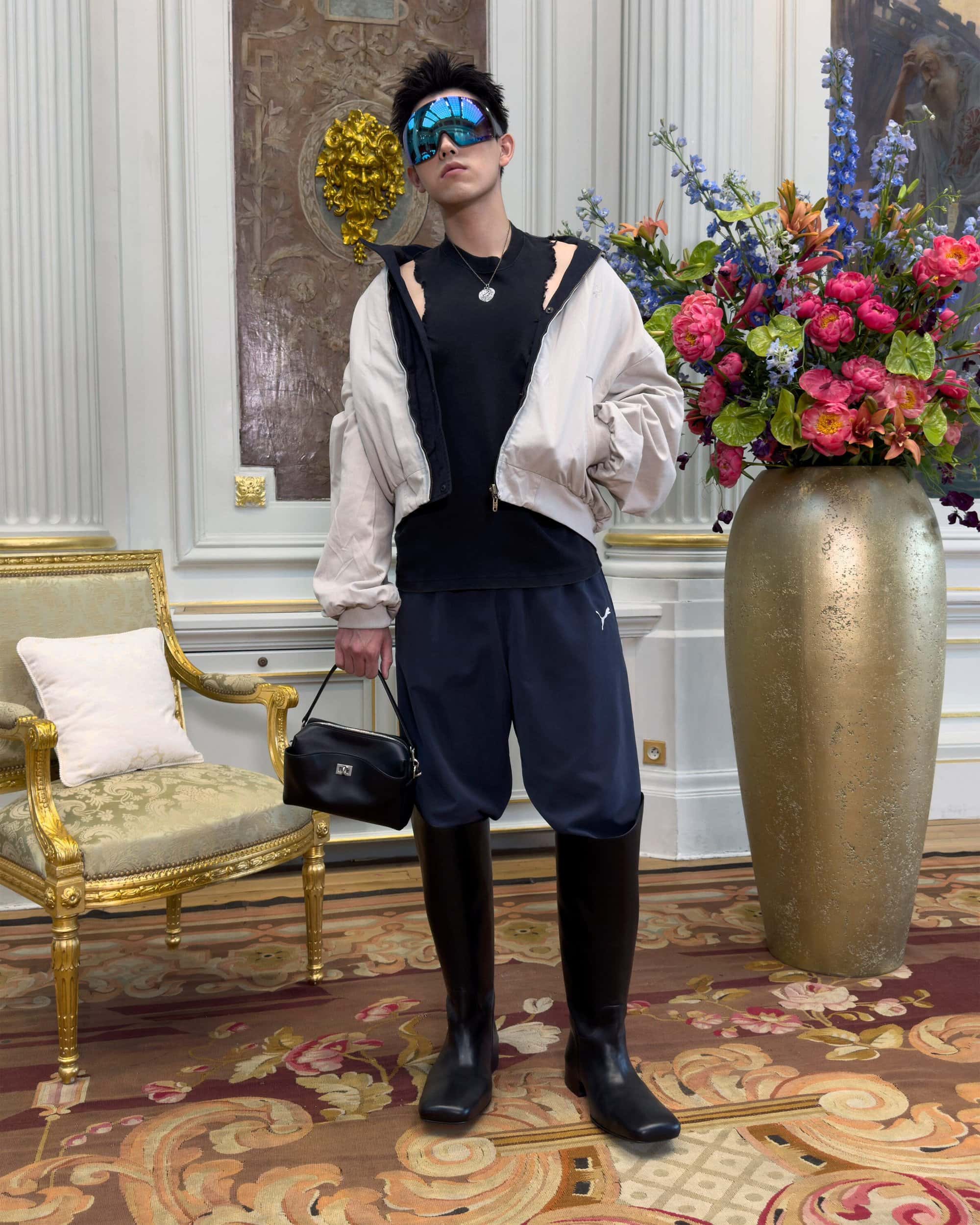 Model wearing oversized sunglasses and tall boots holding a black handbag inside a room with ornate architectural details.