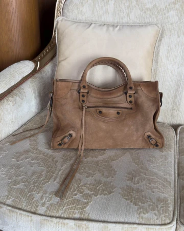 Brown leather handbag with zippered front pocket resting on a patterned upholstered armchair with a cream pillow.
