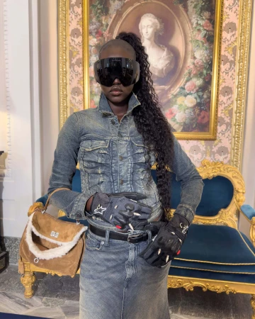 Model in denim jacket and sunglasses holding a suede handbag indoors before a framed oval portrait