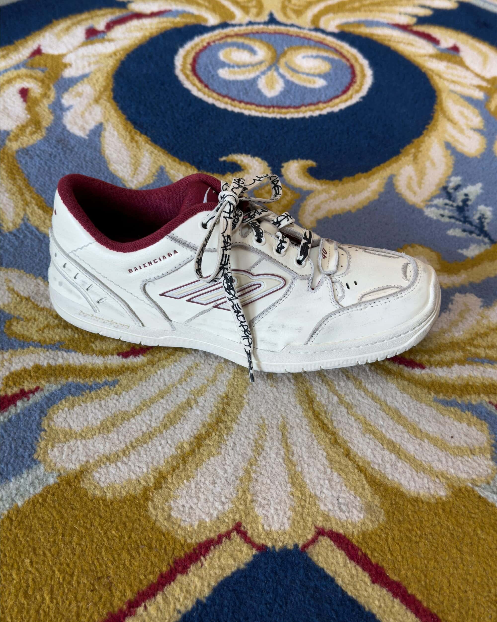 Balenciaga white sneaker with burgundy lining and patterned laces on a blue and yellow patterned carpet.