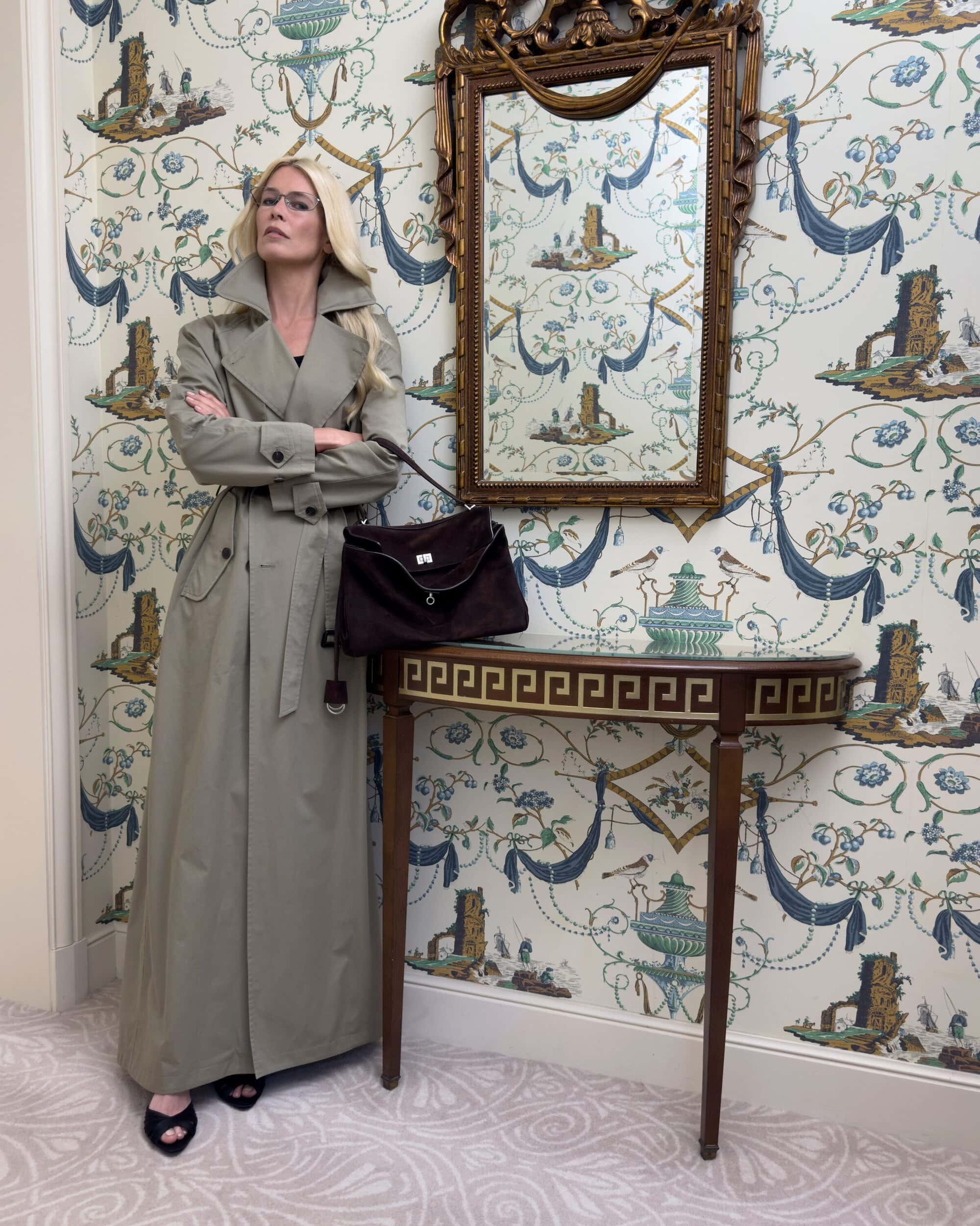 Claudia Schiffer in a long trench coat standing next to a console table against patterned wallpaper for Balenciaga Winter 2025.