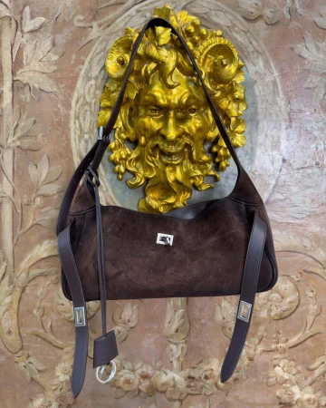 Dark brown suede shoulder bag hanging against an ornate textured wall with a golden relief mask.