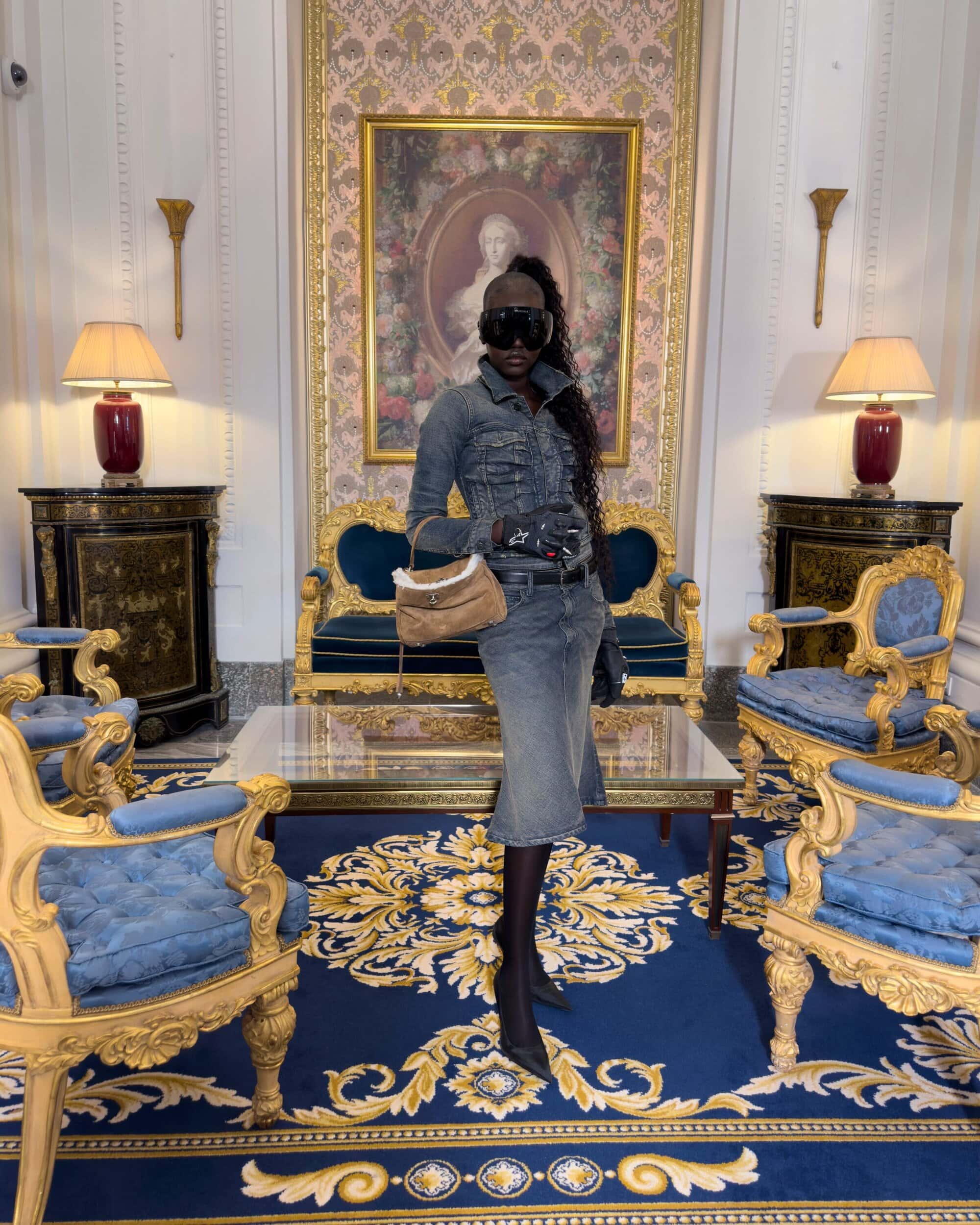 Model wearing a grey denim outfit and sunglasses holding a tan handbag in an ornate room for Balenciaga Winter 2025 ad campaign