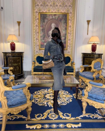Model wearing a grey denim outfit and sunglasses holding a tan handbag in an ornate room for Balenciaga Winter 2025 ad campaign