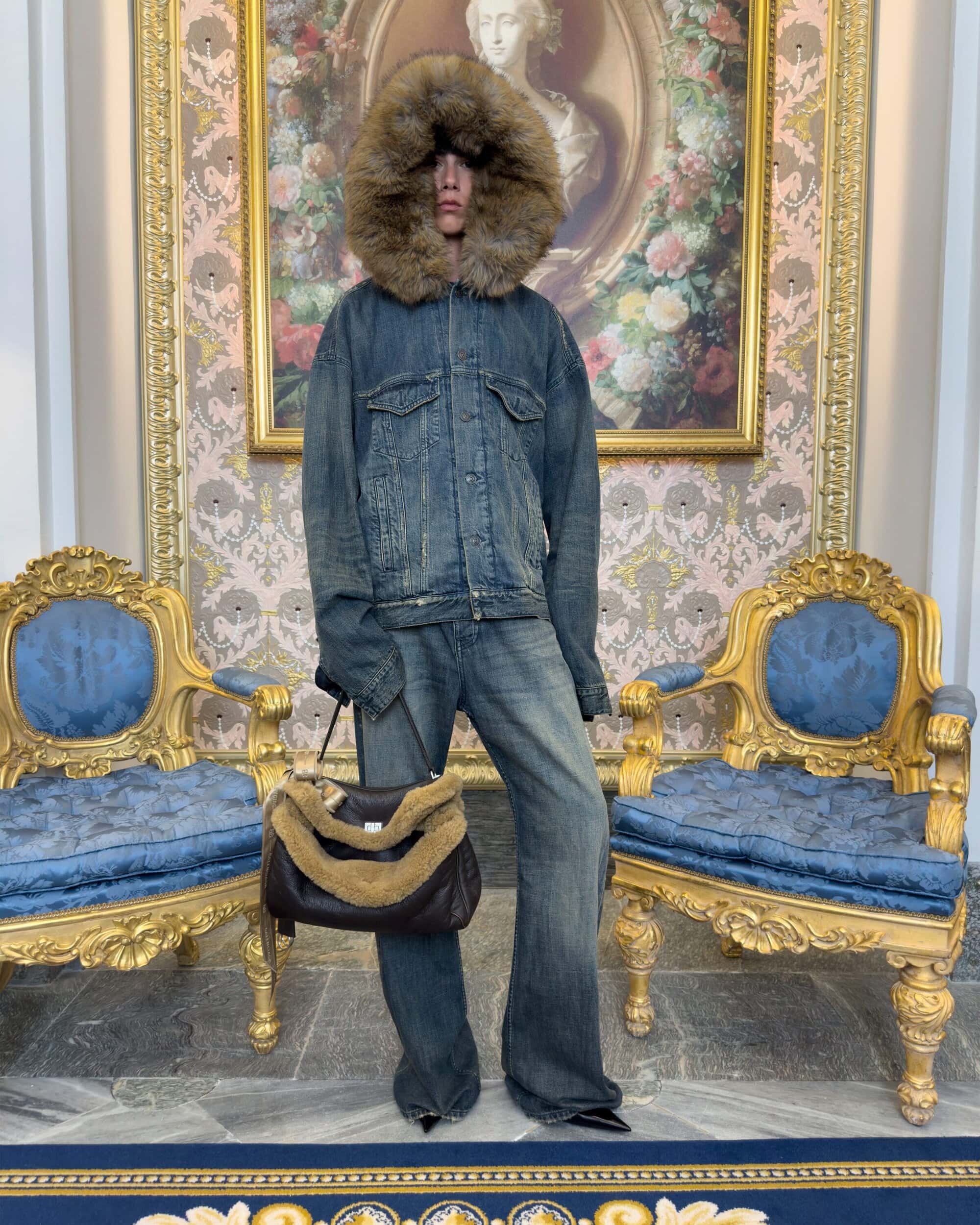 Model standing between two ornate gold chairs wearing an oversized denim jacket with a fur hood and holding a dark handbag.