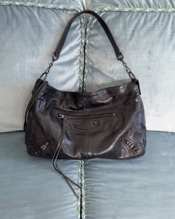 Black leather shoulder handbag with zipper pocket and straps resting on a blue tufted upholstered surface