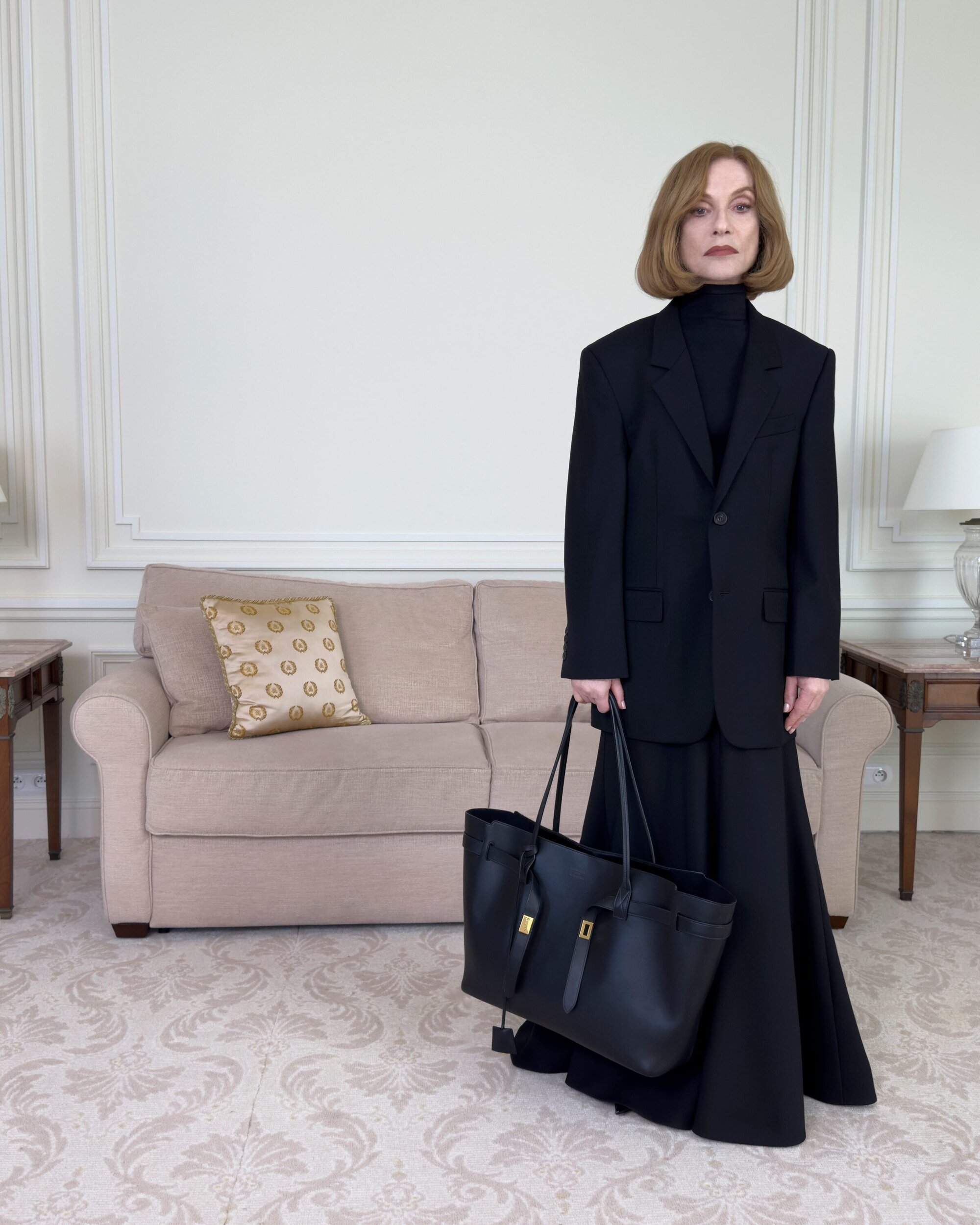 Isabelle Huppert wearing a black oversized suit and holding a black tote bag in a living room for Balenciaga Winter 2025 Ad Campaign