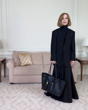 Isabelle Huppert wearing a black oversized suit and holding a black tote bag in a living room for Balenciaga Winter 2025 Ad Campaign
