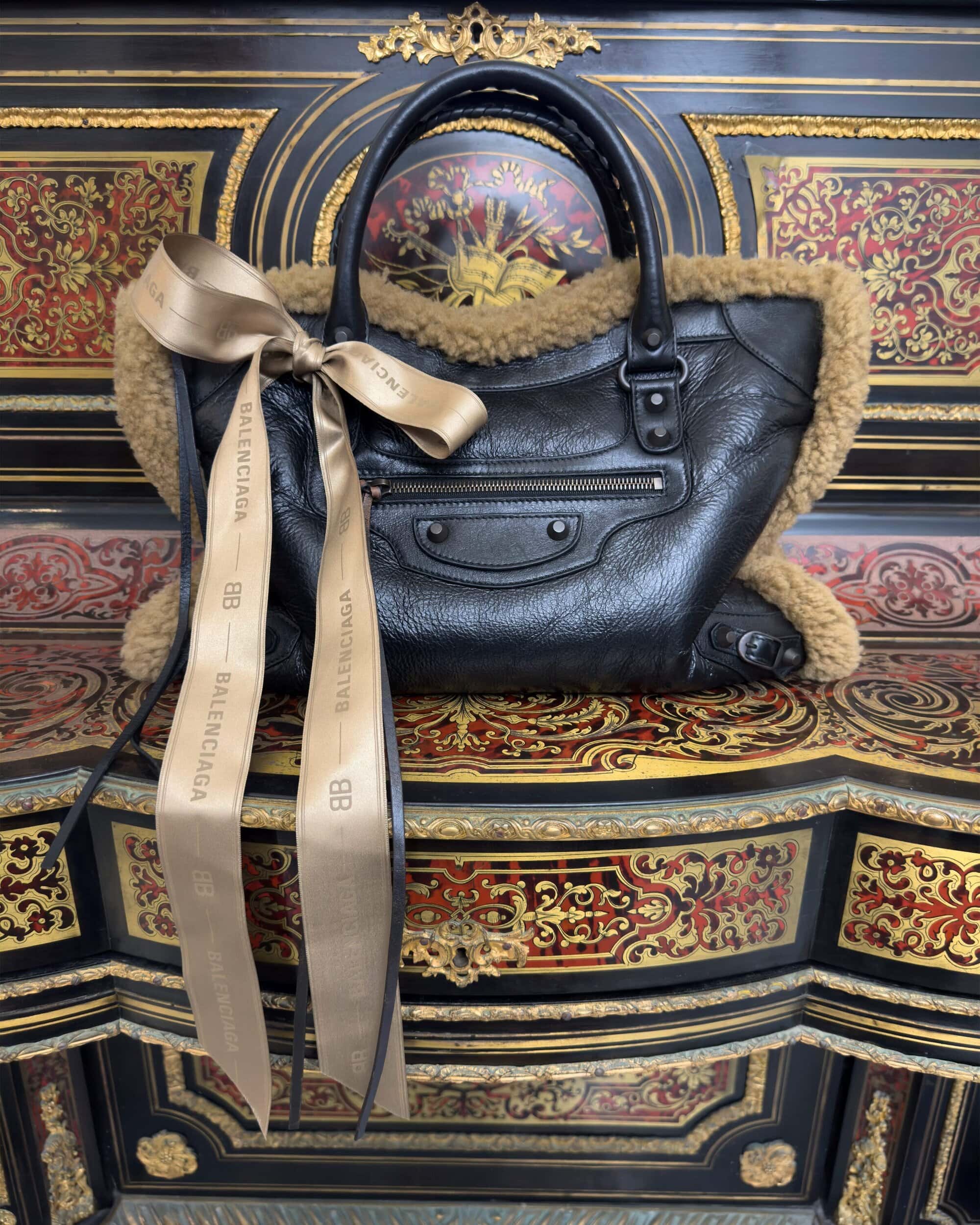 Black leather Balenciaga handbag with shearling trim and tied beige ribbon resting on an ornate antique desk