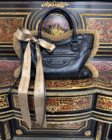 Black leather Balenciaga handbag with shearling trim and tied beige ribbon resting on an ornate antique desk