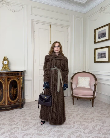 Isabelle Huppert wearing a long brown textured coat holding a dark handbag in an ornate room for Balenciaga Winter 2025 Ad Campaign