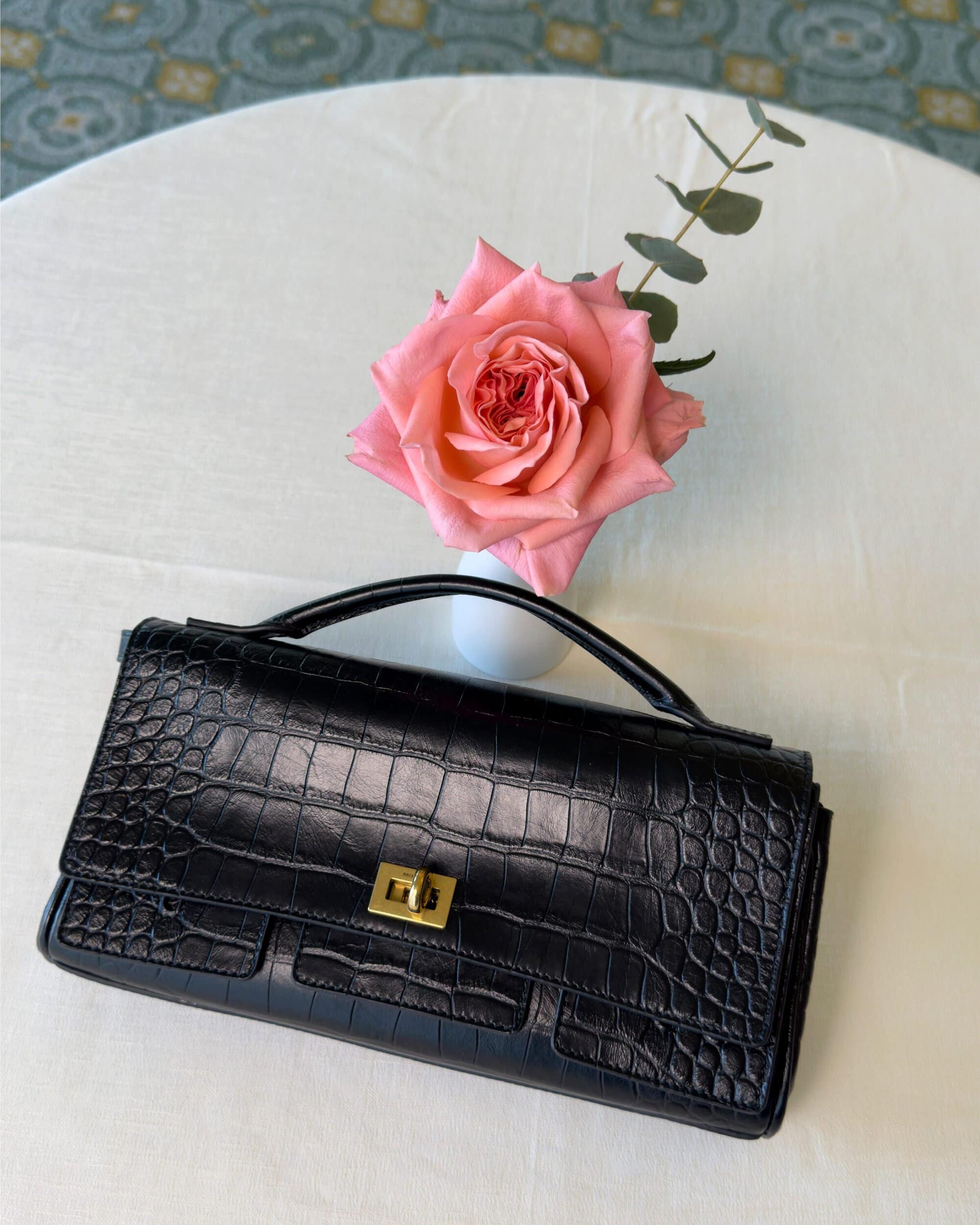 Black crocodile embossed leather handbag on white surface with pink rose in vase