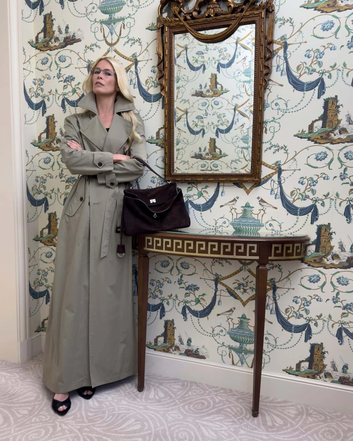 Claudia Schiffer in a long trench coat standing next to a console table against patterned wallpaper for Balenciaga Winter 2025.