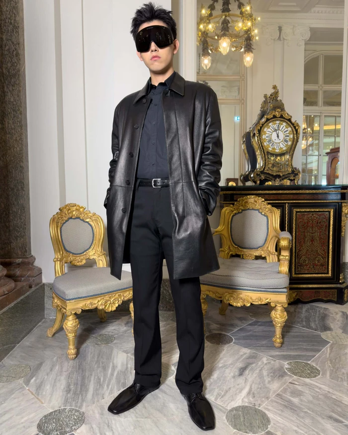 Man wearing a black leather coat, dark shirt, trousers, and sunglasses standing in an ornate room with gilded chairs.