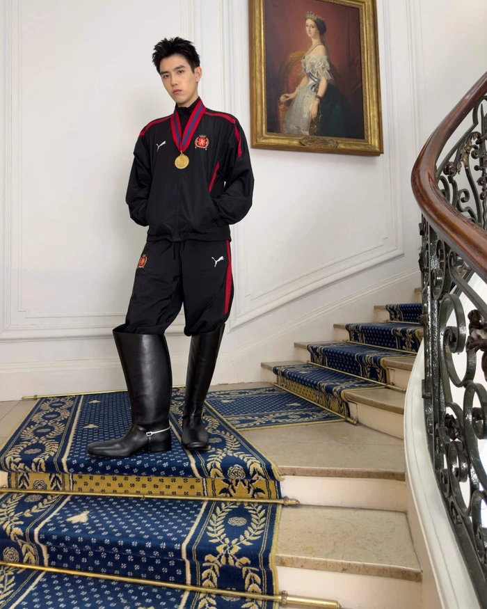 Man wearing a black tracksuit and tall leather boots standing on a grand carpeted staircase beneath a framed portrait