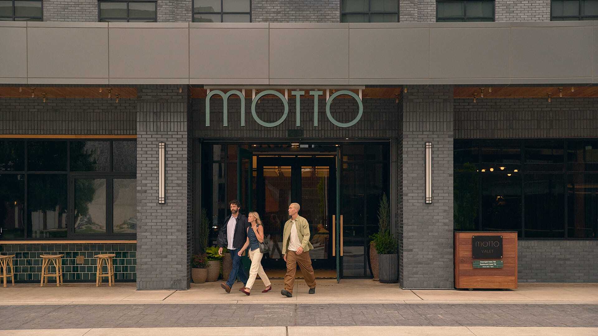 Three people walking out of a Motto hotel entrance beneath a large exterior sign for Motto by Hilton.