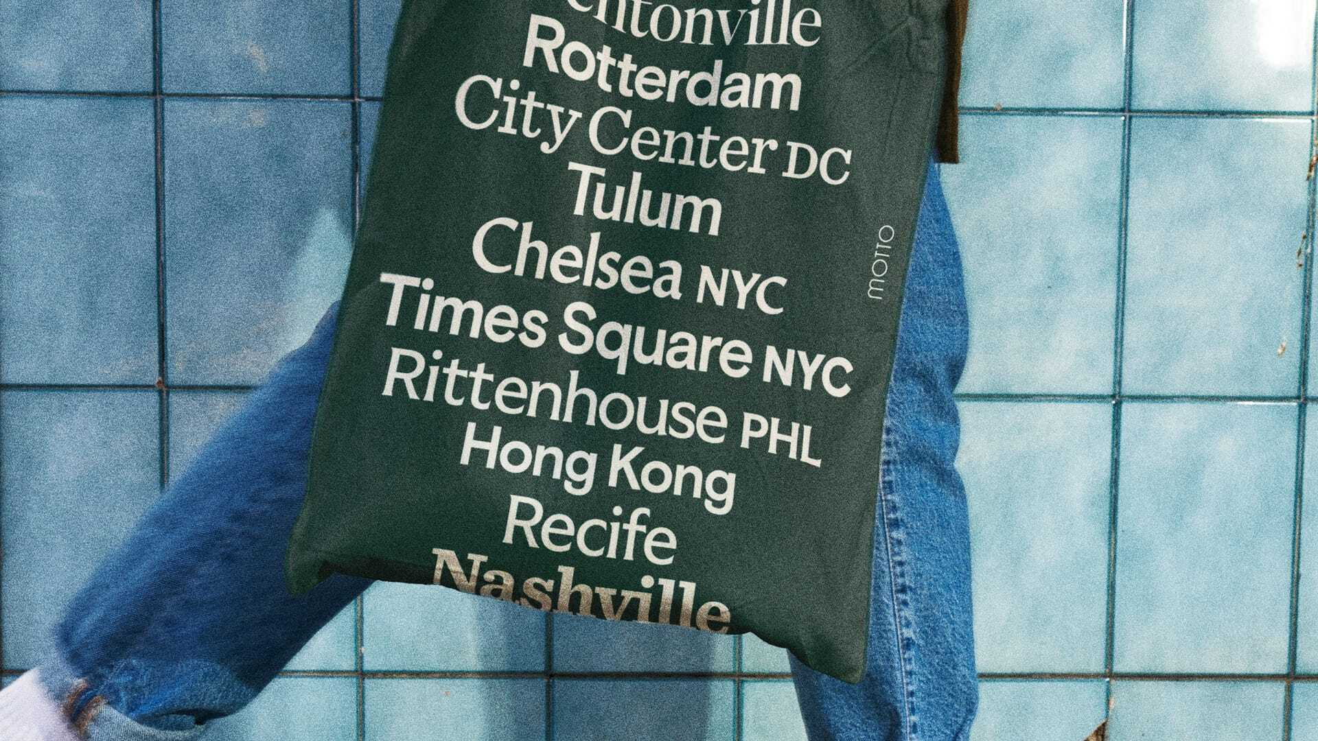 Close-up of a person wearing blue jeans and carrying a dark green tote bag featuring white printed location names against blue tiles