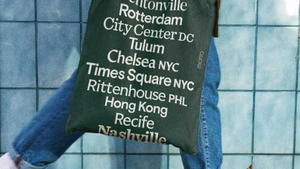 Close-up of a person wearing blue jeans and carrying a dark green tote bag featuring white printed location names against blue tiles
