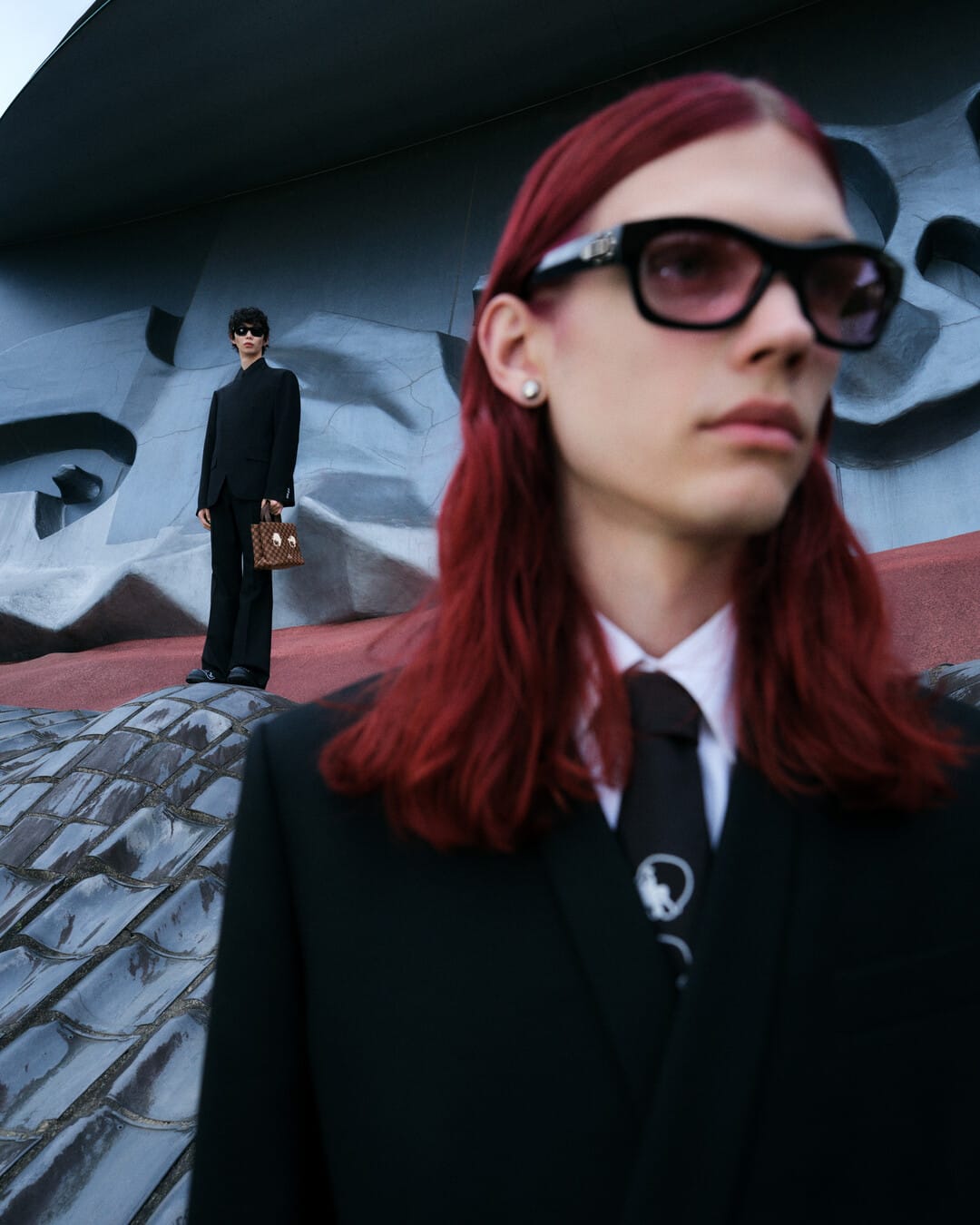 Two male models in black suits and ties outdoors against a sculptural modern architectural background with a handbag.