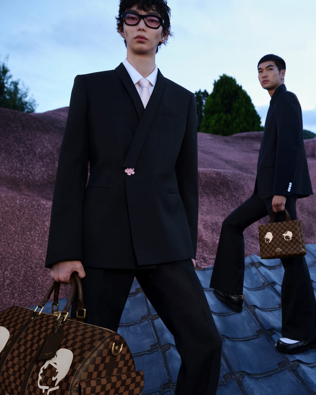 Two men in dark suits holding patterned designer handbags on an undulating purple and grey outdoor terrain.