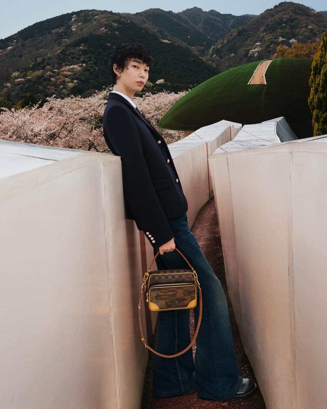 Young man in dark blazer and blue jeans holding a checked handbag, leaning against pale concrete walls outdoors at Yoro Park.