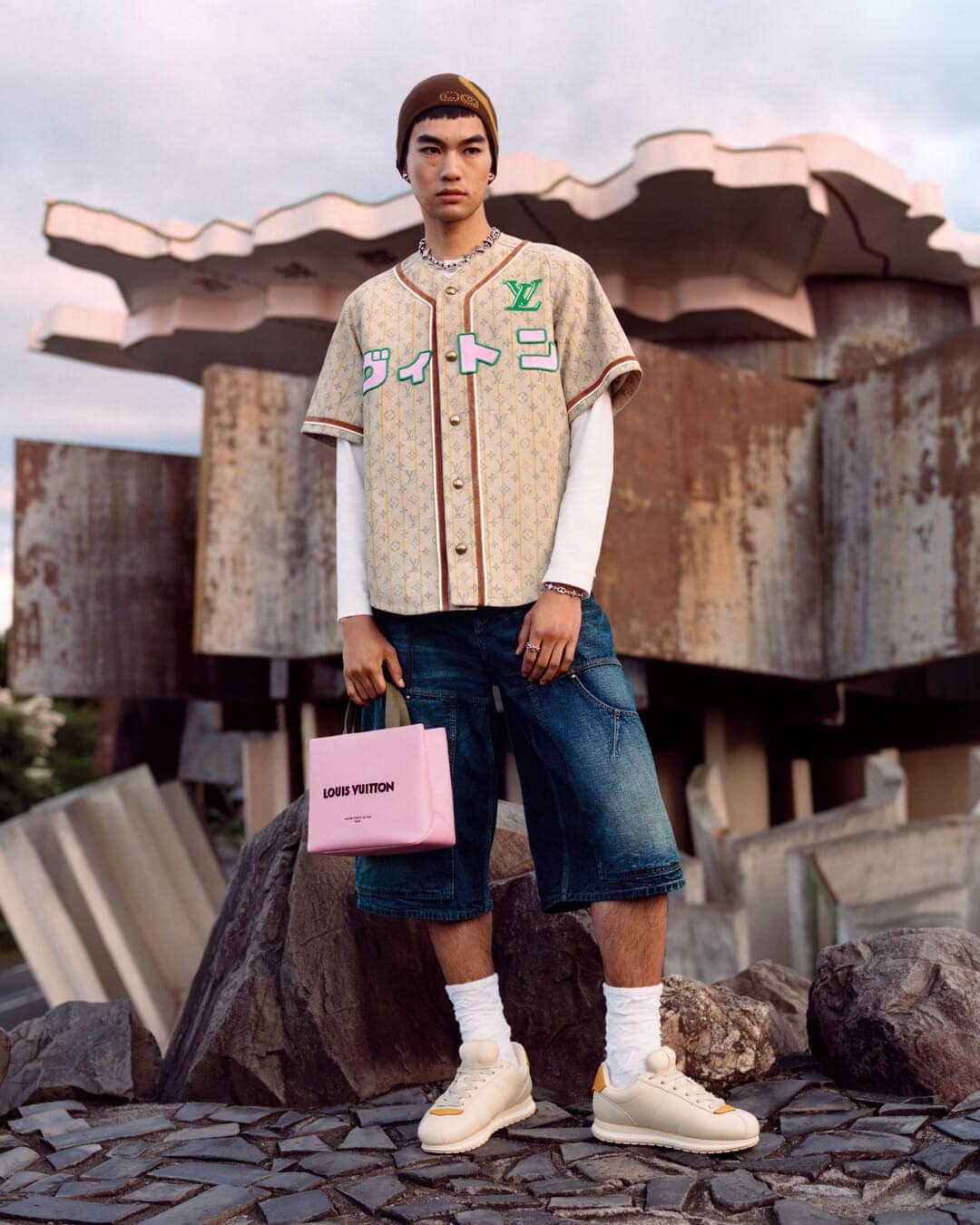 Model in Louis Vuitton monogram baseball jersey holding pink handbag standing outdoors near rocky structures