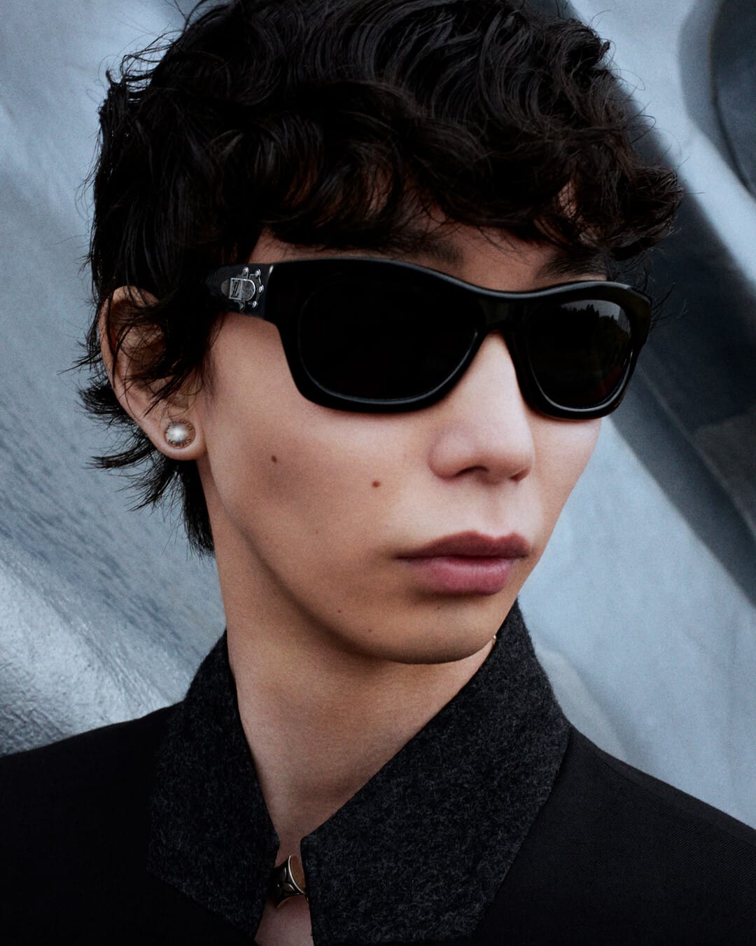 Close-up portrait of a person wearing black sunglasses and a dark collared jacket against an abstract background