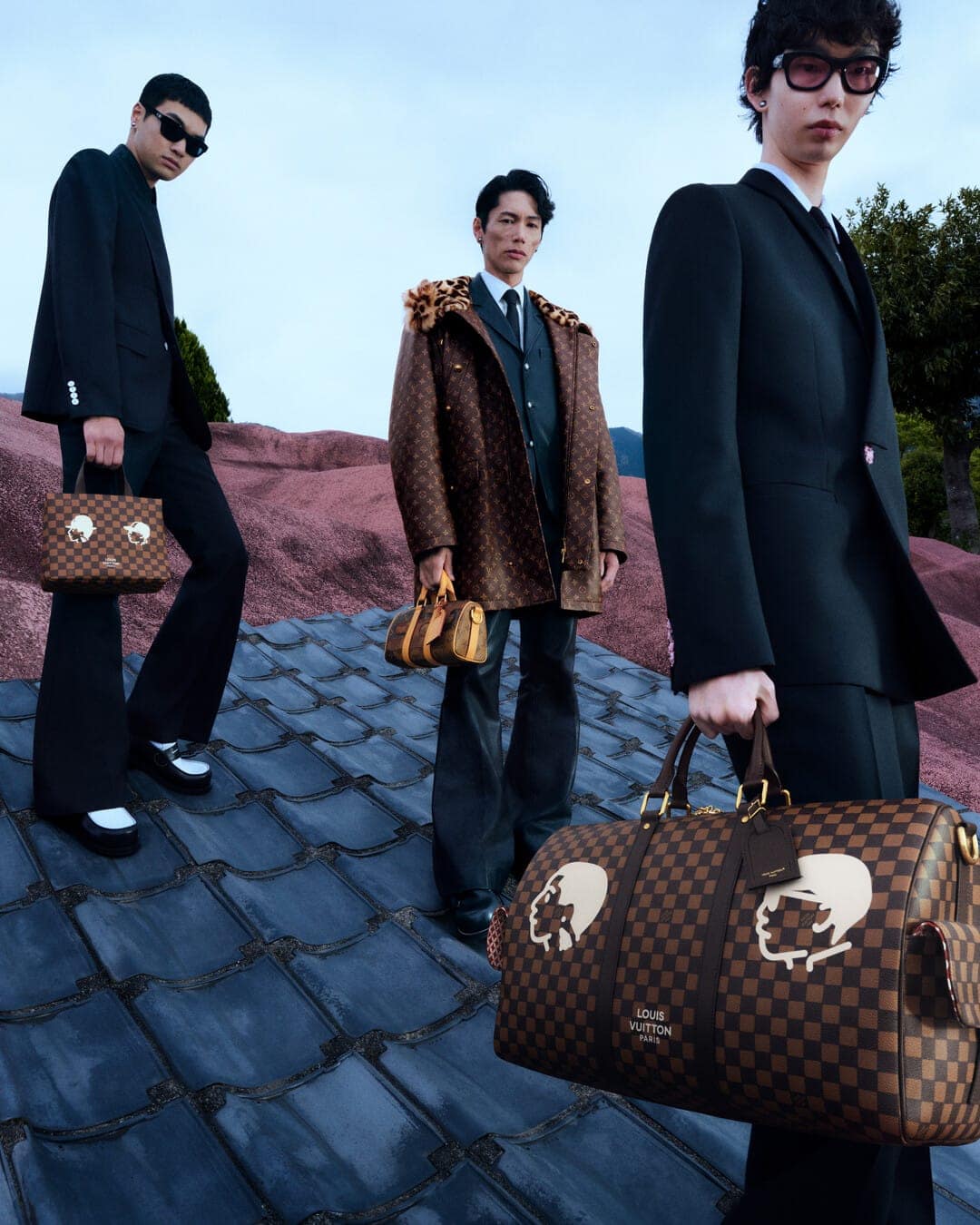 Three male models in suits and monogram coats holding designer duffel bags on a textured pink roof.