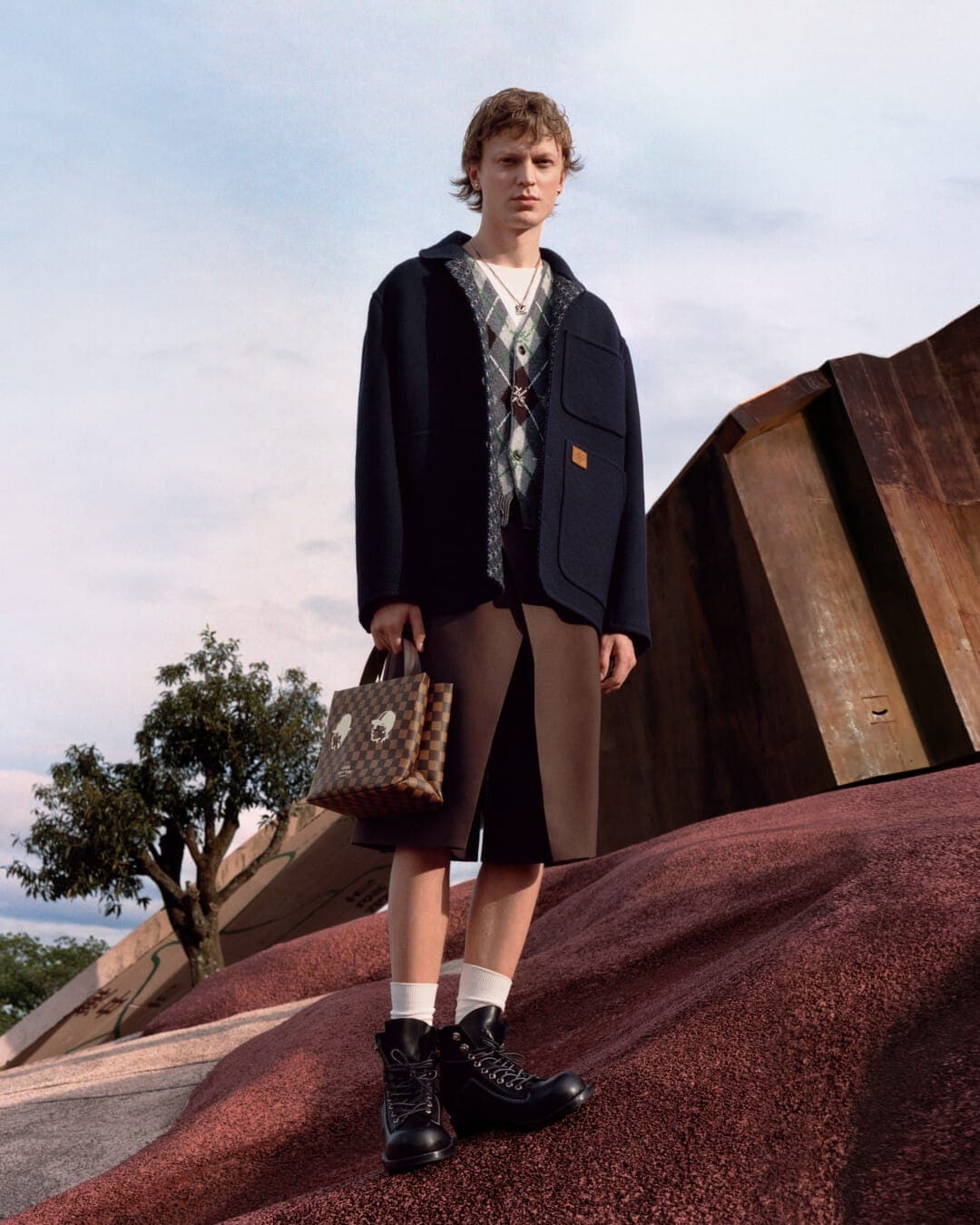 A young male model standing outdoors on a sloped red terrain wearing a dark jacket, argyle vest, shorts, and boots while holding a patterned handbag.