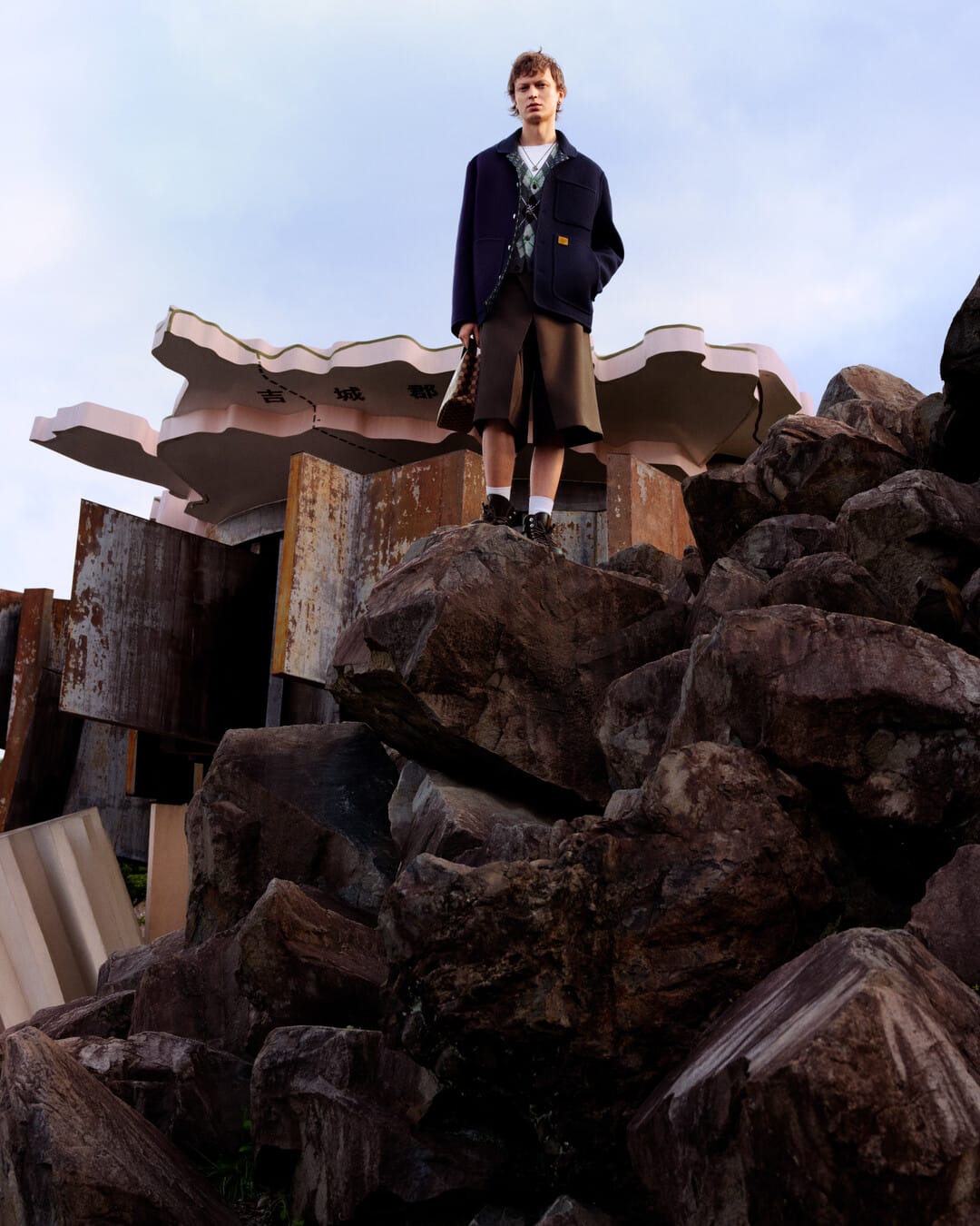 Model standing on rocky outcrop in dark jacket and shorts against pale sky