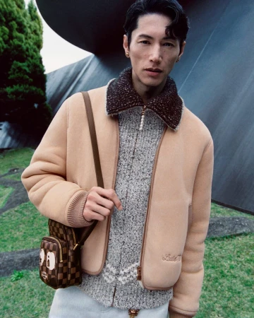 Asian man in camel shearling jacket and checkered crossbody bag standing outdoors with trees and curved architecture.