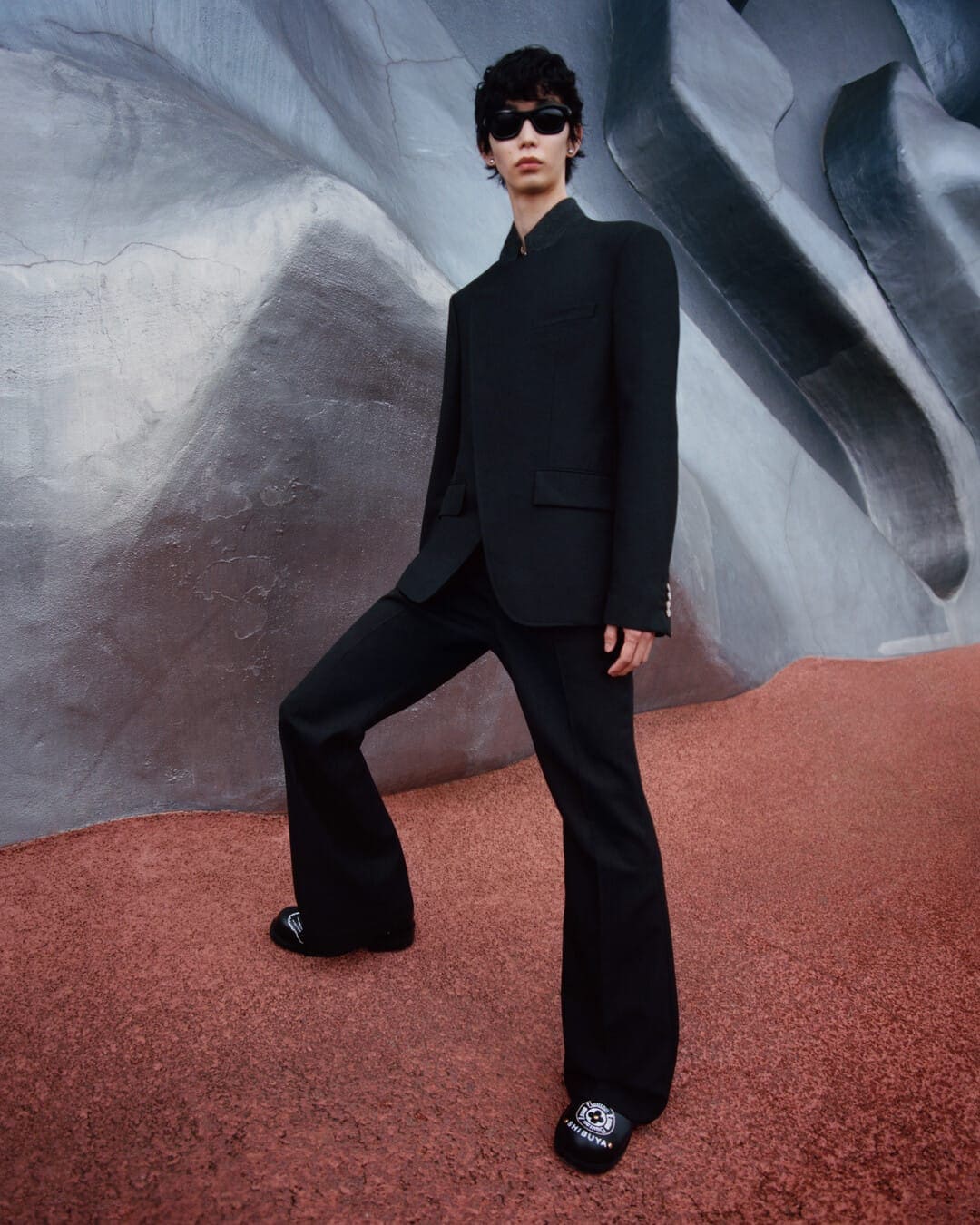 Model wearing a black suit and sunglasses standing on a red textured ground against a sculptural gray stone wall.