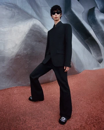 Model wearing a black suit and sunglasses standing on a red textured ground against a sculptural gray stone wall.