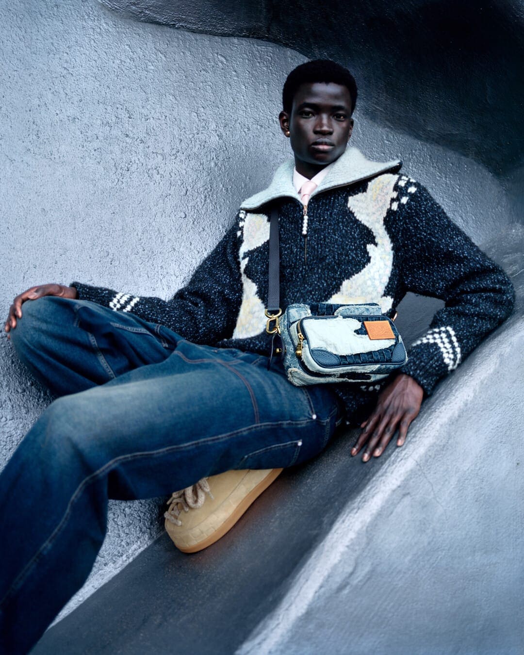 Black man seated in textured grey architectural setting wearing patterned zip sweater, wide jeans, and a crossbody bag.