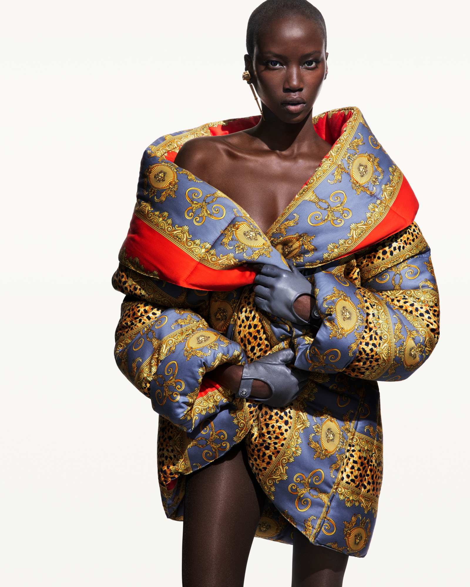 Model wearing a large patterned puffer coat and leather gloves against a plain white background for Versace Fall 2025 campaign.